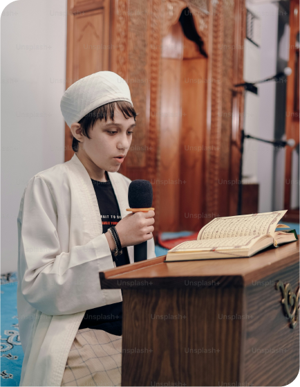 A young boy in traditional attire reading from a book with a microphone in hand, standing at a wooden podium in a mosque